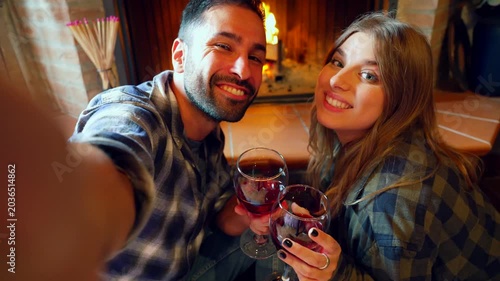 Happy couple taking selfie video by fireplace. Young couple taking a selfie video, drinking red wine, and kissing in front of a fireplace