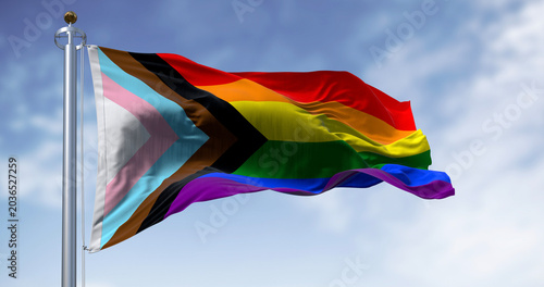 Foto Progress Pride Flag with rainbow stripes and inclusive chevron waving from a silver flagpole against a bright blue sky with light clouds