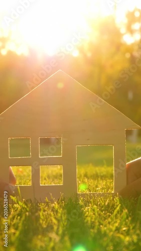 Hands hold wooden house model in sunlight. Bright background creates warm atmosphere. Vertical home concept symbolizes dreams and aspirations. Nature inspires creativity and hope.