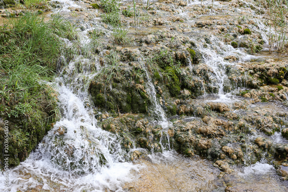 Fototapeta premium Small waterfalls flowing over rocky terrain. Natural cascade landscape in forest environment. Fresh river stream background. Scenic national park attraction outdoors. Water movement and nature detail.