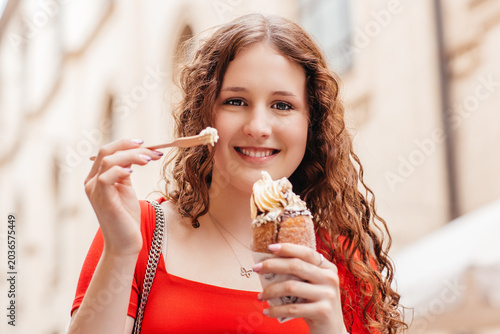 Slika na platnu Smiling young woman eating sweet pastry with cream in city street portrait