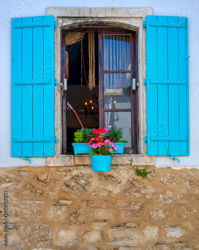View through a brown, half-open window with blue shatters into a dark interior. White facade and rubble stone base in the village of Bellapais, Cyprus.