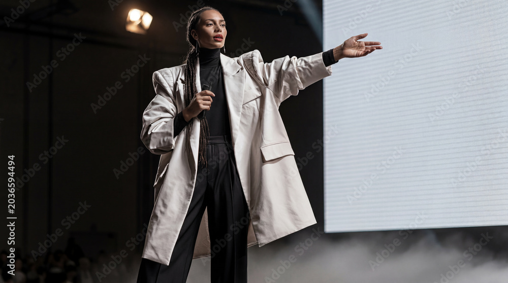 Fototapeta premium Confident young Gen Z businesswoman gesticulating on stage during public speech, female leader presenting startup project at conference hall, career concept.