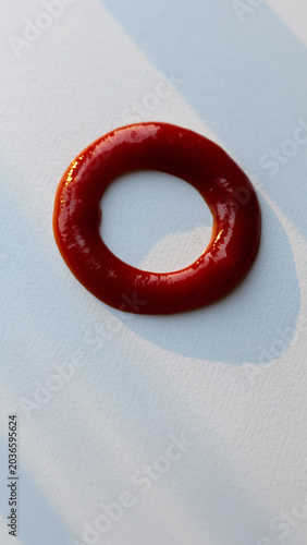 A vibrant, glossy red ring of thick sauce, possibly tomato-based, sits centered on a bright white textured surface under dramatic lighting.