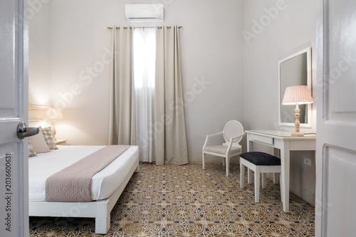 Bright hotel bedroom interior featuring a comfortable bed. Nightstand with a lamp. A dressing table with mirror. And patterned floor tiles. Offering a welcoming space for relaxation and accommodation