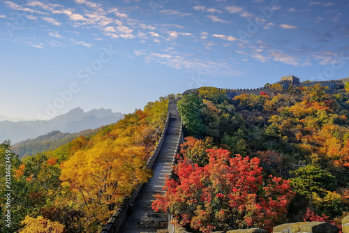 A vibrant autumn scene at the Mutianyu section of the Great Wall of China, with colorful foliage and clear skies. The iconic wall stretches into the distance, surrounded by stunning mountainous views.