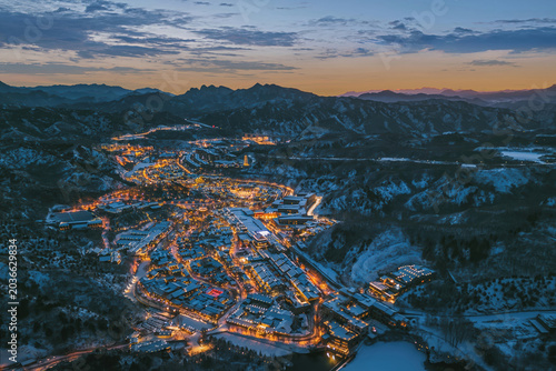 Stunning aerial view of Simatai Great Wall and Gubei Water Town at night, illuminated by warm lights under a twilight sky, surrounded by mountainous terrain.