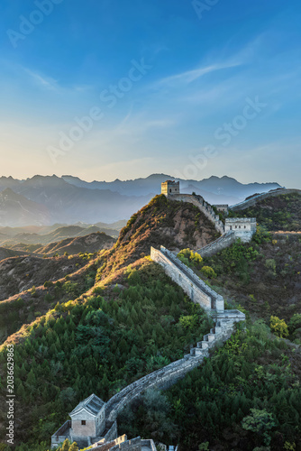 A stunning view of the Jinshanling Great Wall bathed in soft morning light, nestled among the lush green hills of Hebei, China, with misty mountains in the background.
