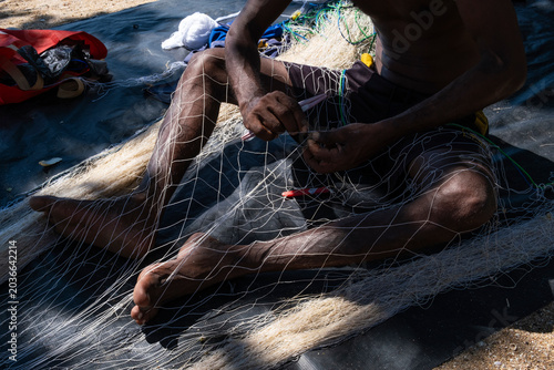 Local Sri Lankan fisherman weaving and repairing nylon fishing net on a sandy beach.