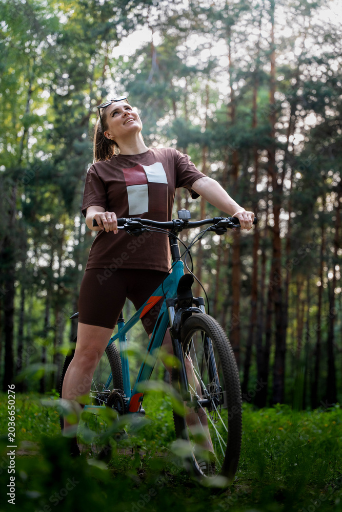 Naklejka premium Woman Standing With Bicycle Looking Skyward Amid Tall Pines, Uplifted Expression, Sunlight Filtering Through Canopy, Sense Of Freedom And Joyful Pause