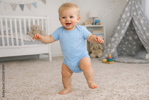 Joyful baby in light blue onesie walking with arms outstretched and smiling at the camera on plush beige carpet with wooden crib featuring white railings and colorful pennant banner.