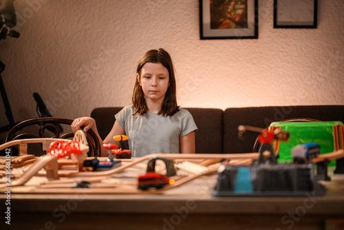Young girl planning her wooden toy train route at home table illuminated by warm evening light