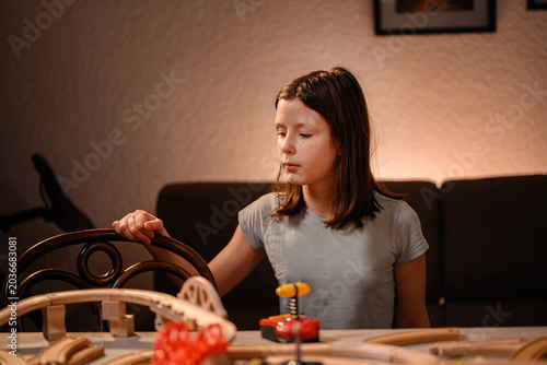 Focused young girl sitting at home table looking at wooden toy railway tracks during cozy evening