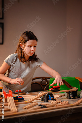 Active young girl pushing red toy train on wooden tracks building railway system in cozy room