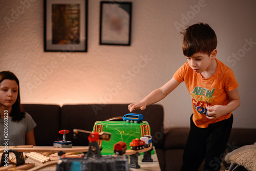 Concentrated boy placing toy train on bridge while sister watches during evening indoor playtime