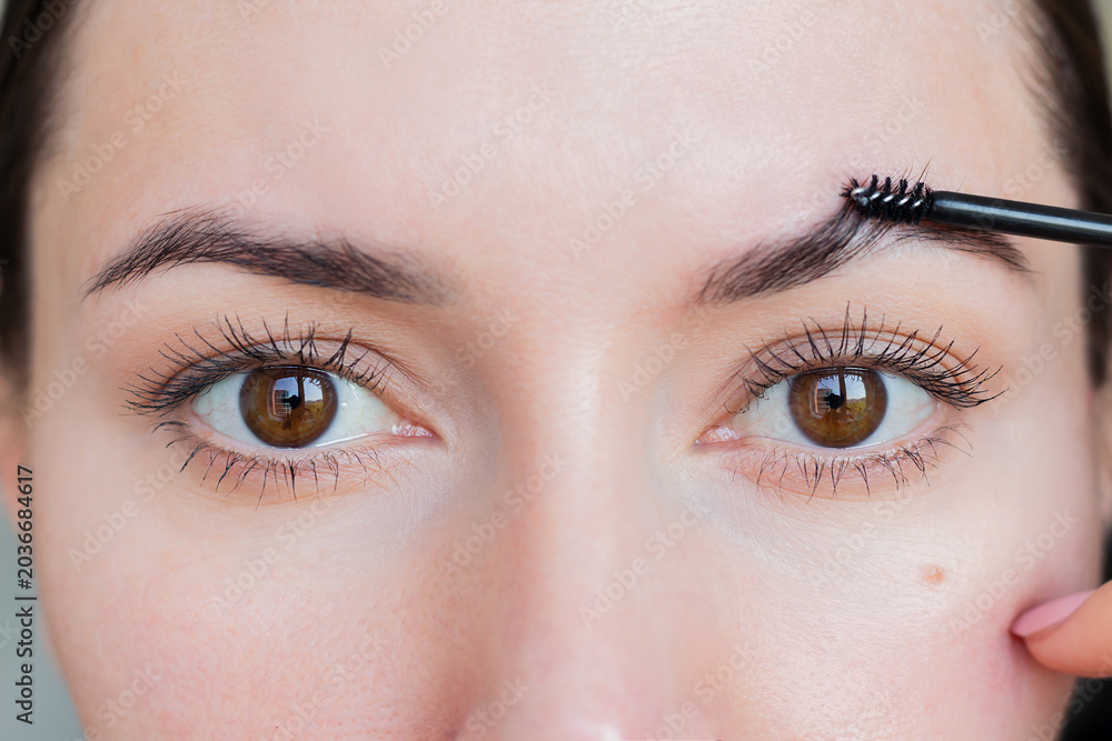 Naklejka premium Young woman combing her eyebrows with a brush, making a beautiful shape. Eyebrow care. Eyebrow styling with gel for fixation.