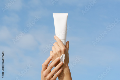 Hands applying white sunscreen from a blank mockup tube against a clear blue sky. Bright, natural lighting with a minimal, clean aesthetic. Perfect for skincare, summer, and sun protection concepts