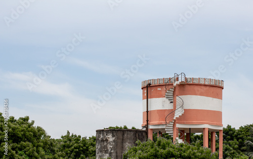 Elevated water storage tank with blue railings on stilts, standing against green trees and cloudy sky, a common rural water supply infrastructure.