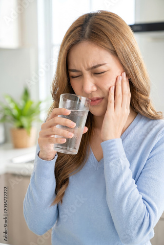 Young Asian woman suffering from sudden tooth sensitivity while trying to drink a glass of ice water at home.
