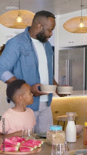 Vertical video: Mom placing watermelon at counter dad in denim holding bowls serving breakfast