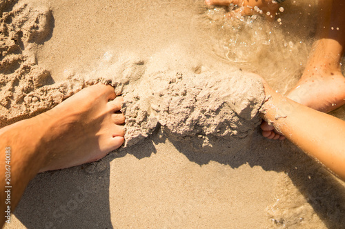 Bild auf Leinwand A close-up action shot of a parent and child's feet playing together and burying each other under the wet sand near the water