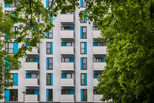 Modern residential building facade with white balconies and bright blue decorative wall panels framed by green summer tree branches.