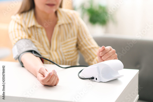 Senior woman measuring blood pressure at white table indoors, selective focus
