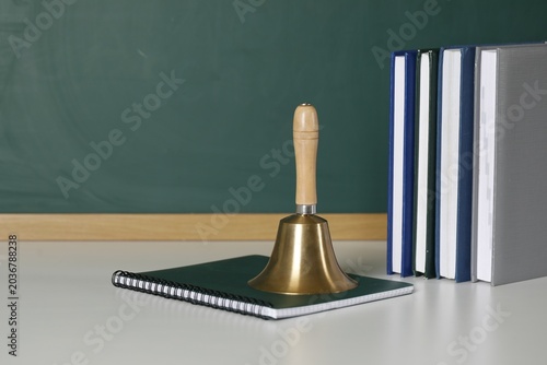 Фотография School golden bell, copybook and notebooks on white table near green chalkboard