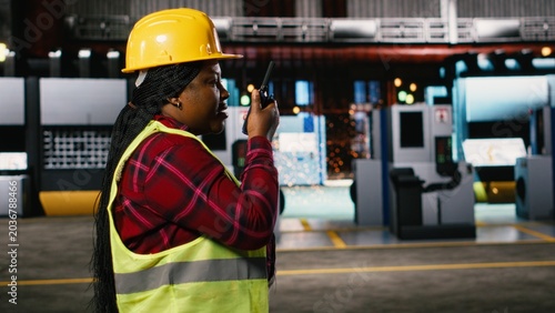 Black woman engineer speaking into portable walkie-talkie device, walking through warehouse and monitoring heavy equipment, Large-scale industrial facility highlights production line.