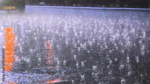 Close view of wet asphalt road surface during heavy rain with colorful orange and red light reflection, rain drop splash pattern and passing car wheel visible
