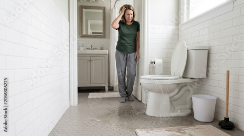 Overflowing toilet in bathroom with concerned woman looking at mess on floor. Toilet overflow creates water damage, causing stress and frustration.