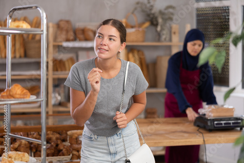 Foto Pleased young girl chooses some bakery goods while Muslim seller works in backgr