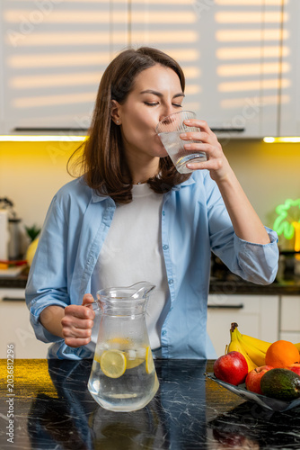 Fotografia Young woman pours cold lemon water into glass then drinks to quench thirst with happy smile in clean kitchen post workout