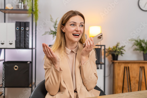 Middle-aged happy businesswoman at home office takes smartphone call with good news turns on speaker laughs and says thanks with joy. Freelancer girl at table smiles listens hands free plans response