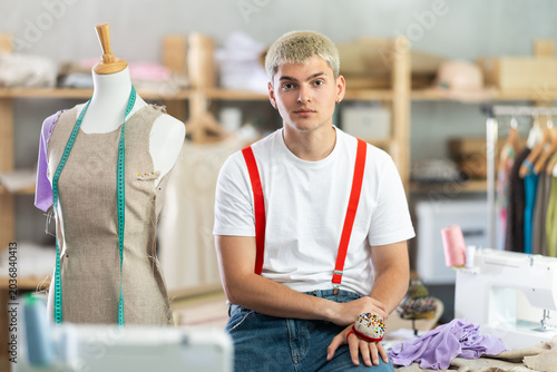 Фотография Content young male tailor half-sitting on table at his workplace in sewing studi