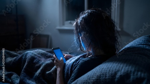 Distressed female holding mobile phone in dark bed late at night unable to sleep due to stress and screen glare