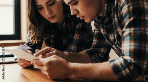 Young man and woman analyze document together. Couple in plaid shirts focus on paper  discussing details at table. Focused team reviewing project at cafe or home. Collaborative work in casual setting.