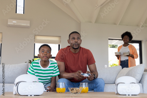 Quadro su tela African American family father and child sitting on sofa playing with controller