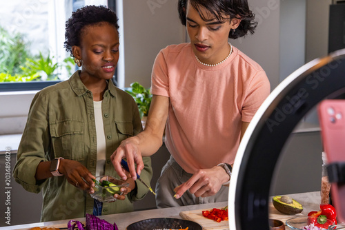 Diverse friends in olive and peach tops making salad at kitchen counter, recording with ring light