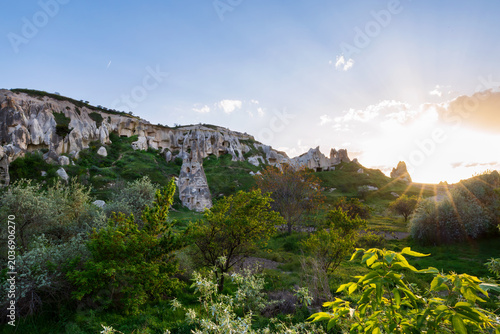 Scenic sunset view of Göreme valley in Cappadocia, Türkiye, with fairy chimney rock formations, ancient rock-cut caves and lush spring greenery illuminated by warm evening light.