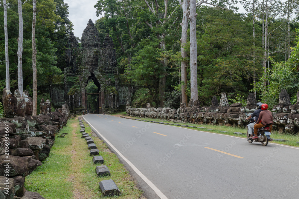 Fototapeta premium Two people ride a motorcycle past the ancient stone statues lining the road to the Angkor Thom North Gate, Siem Rep, Cambodia.