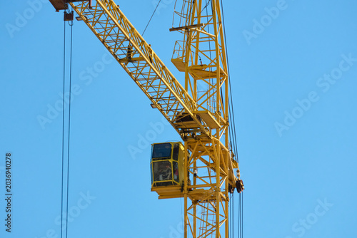 Two tall cranes stand in an urban area during the day. The cranes are positioned against a clear blue sky