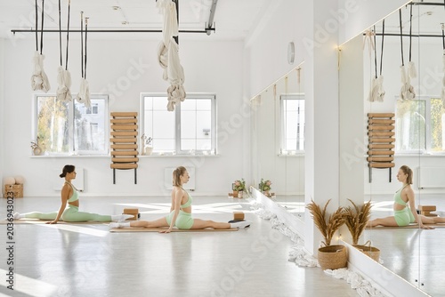 In a spacious yoga studio filled with natural light, two women perform stretches on mats. The room features large mirrors, calm decor, and hanging ropes