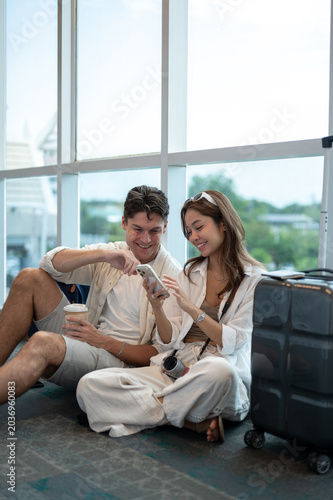 Asian woman girlfriend smiling at phone with white man boyfriend an luggage sits on floor in airport