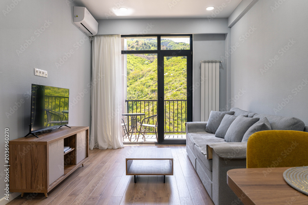Fototapeta premium Modern living room with large window and balcony overlooking green hills, cozy apartment interior