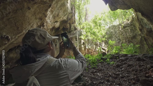 Behind the shot of an Asian man adventurer using a smartphone to take photos towards a cave skylight with natural light.