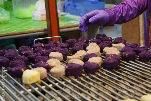 Close up of a street vendor hand arranging colorful purple and yellow sweet potato grilled cakes on a metal wire rack at a local Asian night market stall.