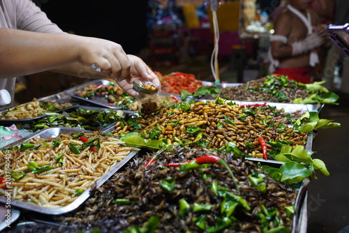 A street food vendor serving various deep-fried edible insects mixed with Thai herbs, presented as a high-protein alternative food source at a local night market.