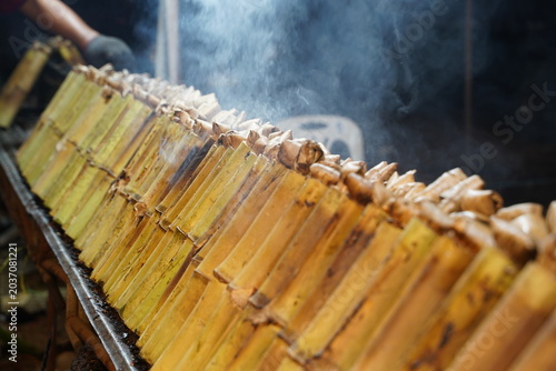 A row of bamboo tubes filling with sweet coconut sticky rice (Khao Lam) being roasted over charcoal grill with smoke rising up at a street food stall.