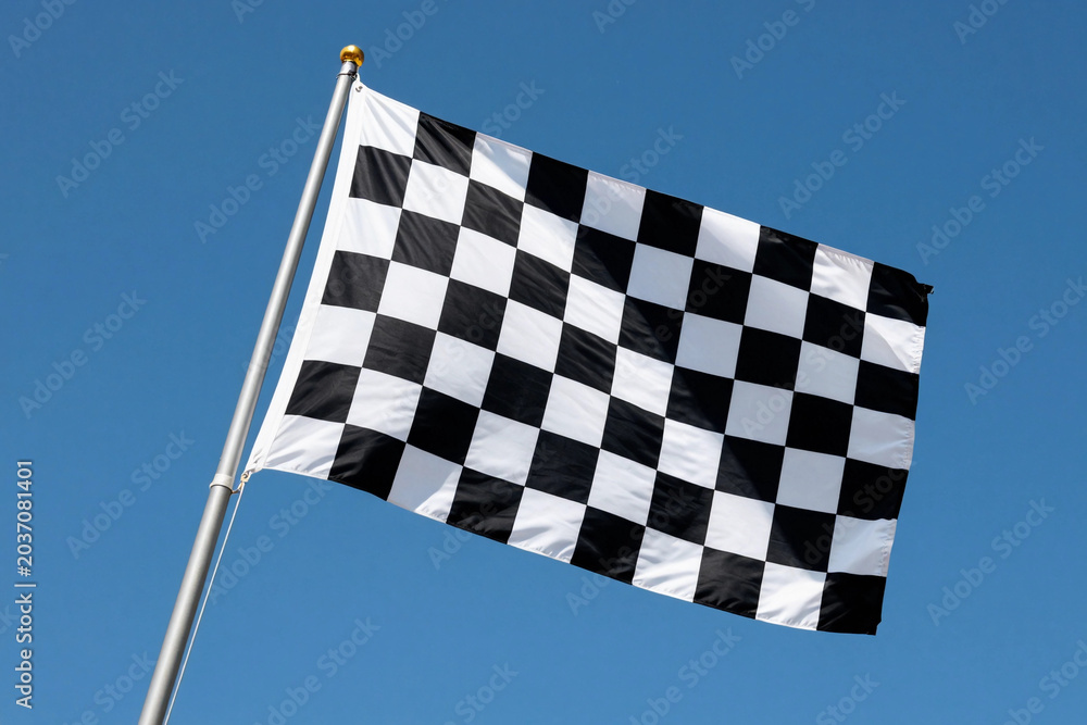 Obraz premium Black and white checkered flag flutters against a clear blue sky. Wind carries the flag with dynamic motion.